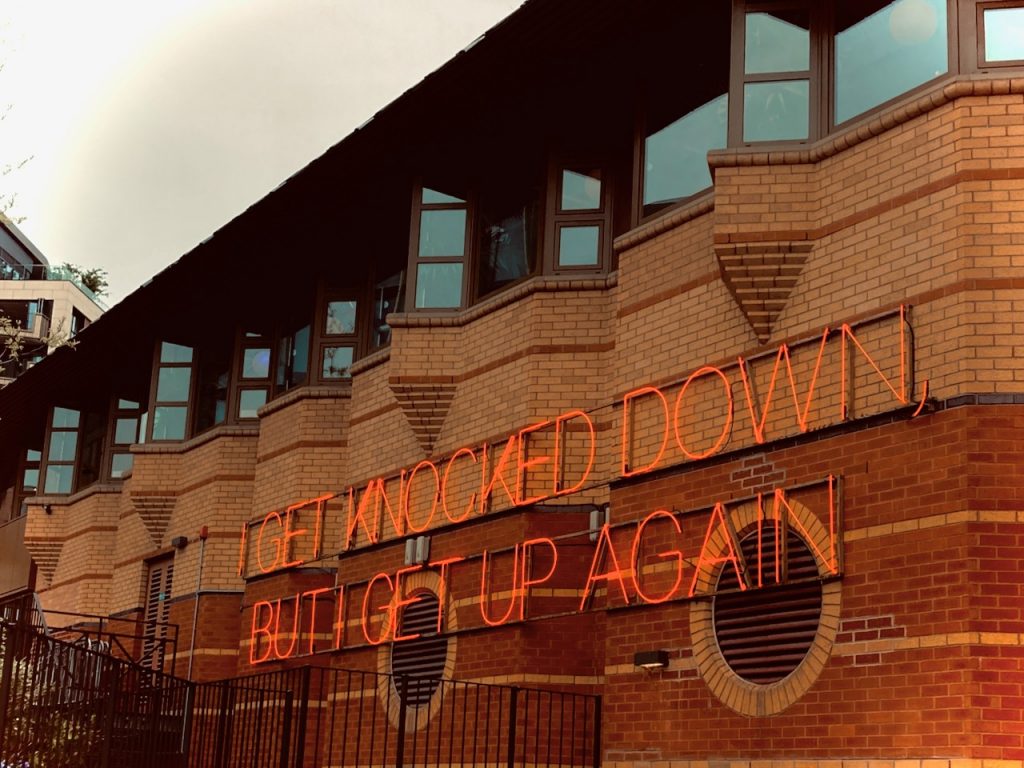 A neon sign on the side of a wall, lyrics from Chumbawamba's song 'Tubthumping'- 'I GET KNOCKED DOWN, BUT I GET UP AGAIN'