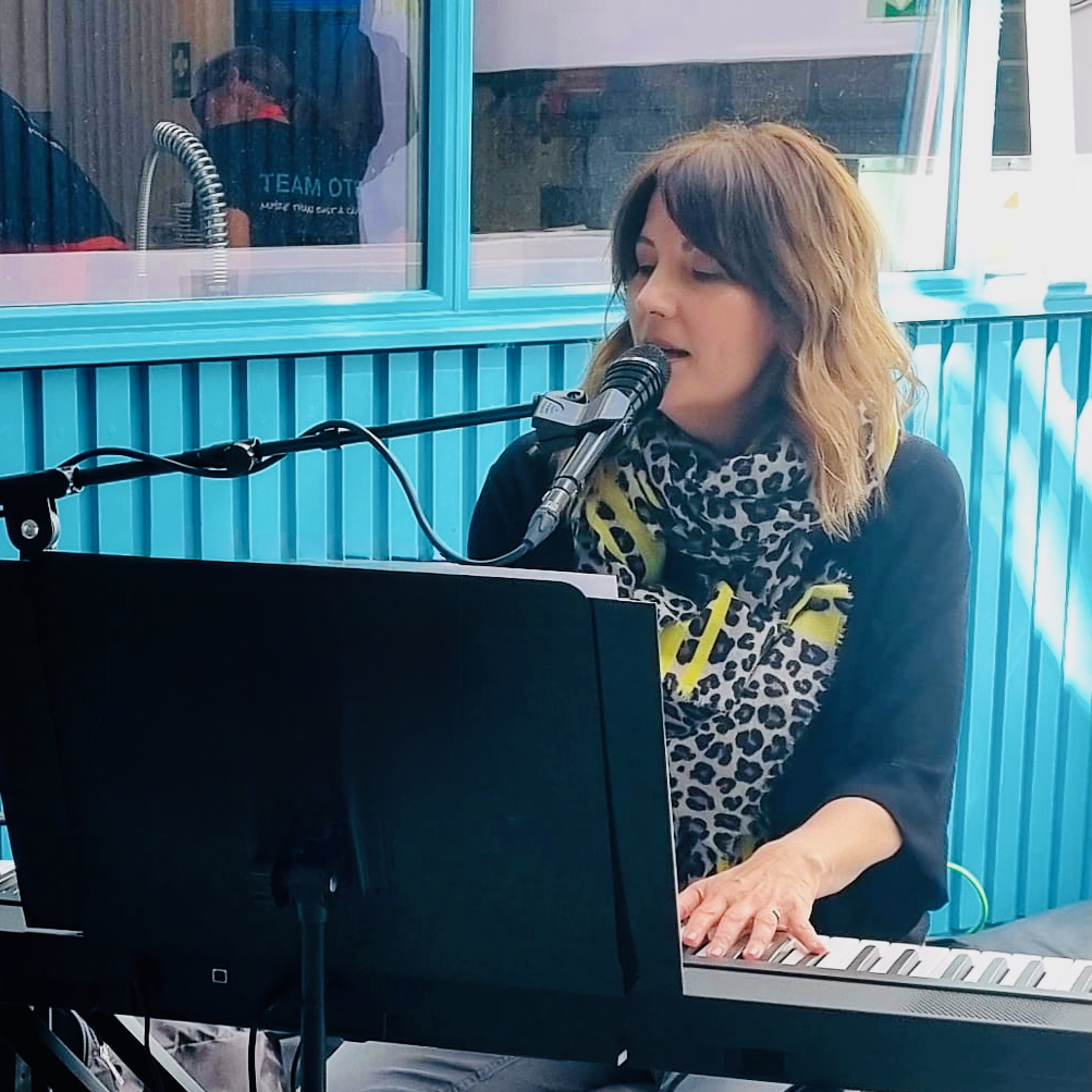 Andrea Mann - a white woman with midlength brown hair - sings and plays a piano keyboard