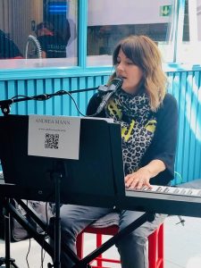 Andrea Mann - a white woman with midlength brown hair - sings and plays a piano keyboard