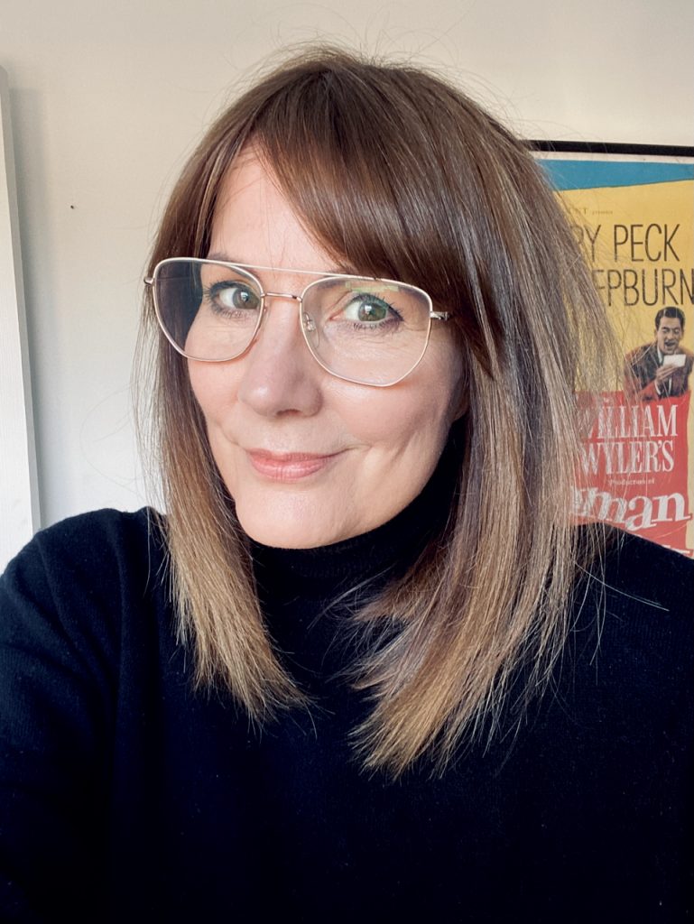 Andrea Mann: a white woman with medium length brown hair, wearing glasses and a dark turtleneck jumper, with a Roman Holiday film poster behind her