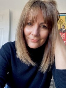 British screenwriter Andrea Mann: a white woman with medium length brown hair, wearing a dark turtleneck jumper, with a Roman Holiday film poster behind her
