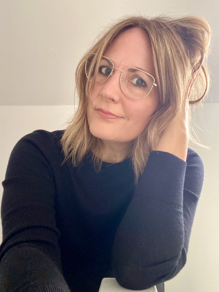 Andrea Mann: a white woman with medium length bronde hair, wearing gold-framed glasses and a dark jumper